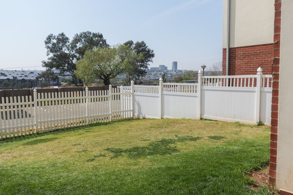 A private rear garden area featuring a well-maintained vinyl picket fence, brick exterior wall, and a grassy lawn with distant urban views.