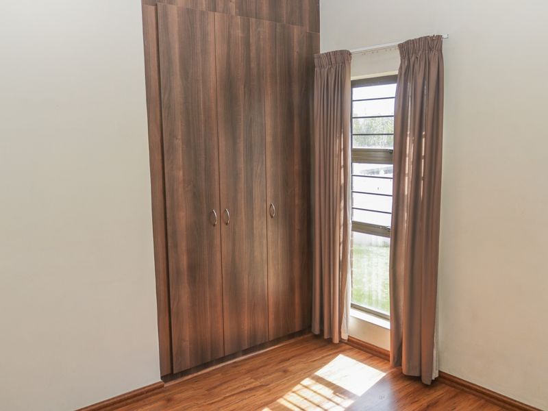 A bright, empty bedroom featuring modern wood-finish built-in cupboards, laminate flooring, and a window with security bars.