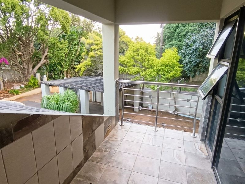 A covered balcony featuring tiled flooring and a stainless steel balustrade, offering an elevated view of the surrounding garden and property grounds.
