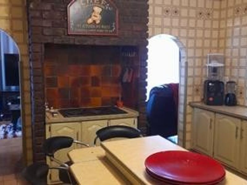 A dated kitchen featuring a prominent brick fireplace surround, built-in hob, and traditional cabinetry with arched architectural details connecting to adjacent living spaces.