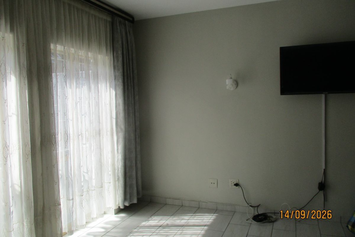 A sparsely furnished living area featuring tiled flooring, wall-mounted lighting, and large windows with curtain tracks, requiring minor cosmetic updates.