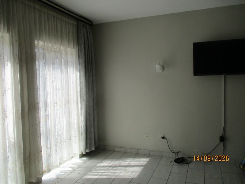 A sparsely furnished living area featuring tiled flooring, wall-mounted lighting, and large windows with curtain tracks, requiring minor cosmetic updates.