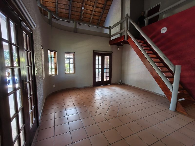 Spacious living area featuring a double-volume thatched ceiling, exposed timber staircase, terracotta tiled flooring, and multiple sets of french doors providing natural light.