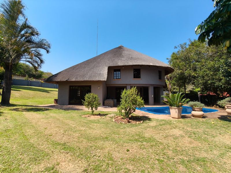 Rear view of a property featuring a traditional thatch roof, a swimming pool, and a paved patio area set within a spacious garden.