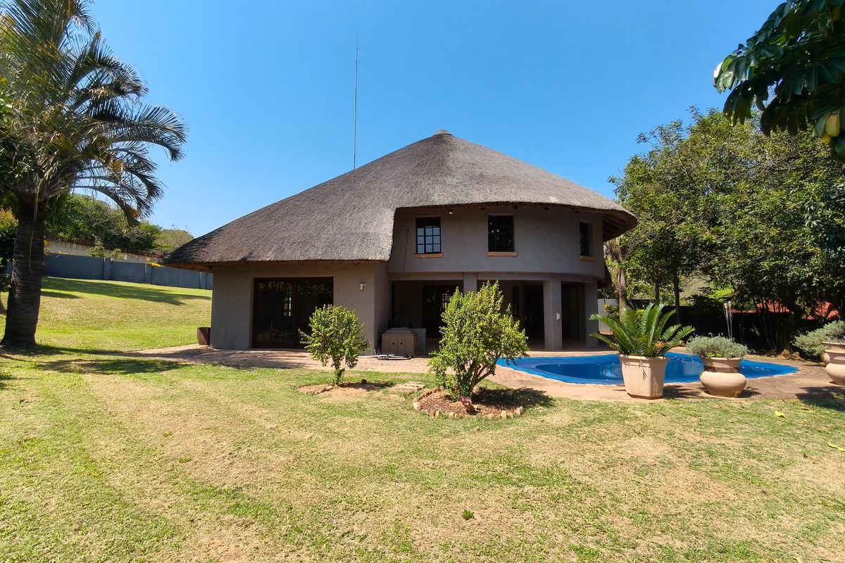 Rear view of a property featuring a traditional thatch roof, a swimming pool, and a paved patio area set within a spacious garden.