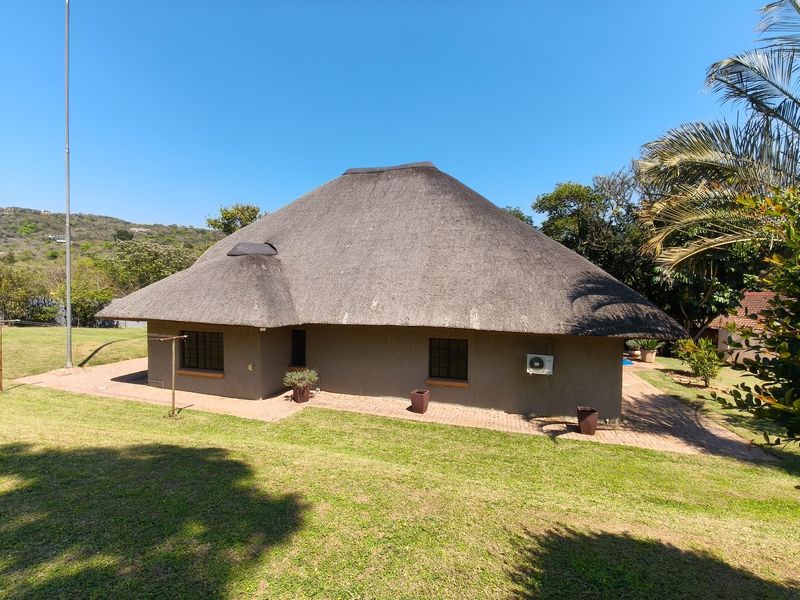 Rear view of a single-story house featuring a traditional thatch roof, brick paved patio area, and surrounding lawn.
