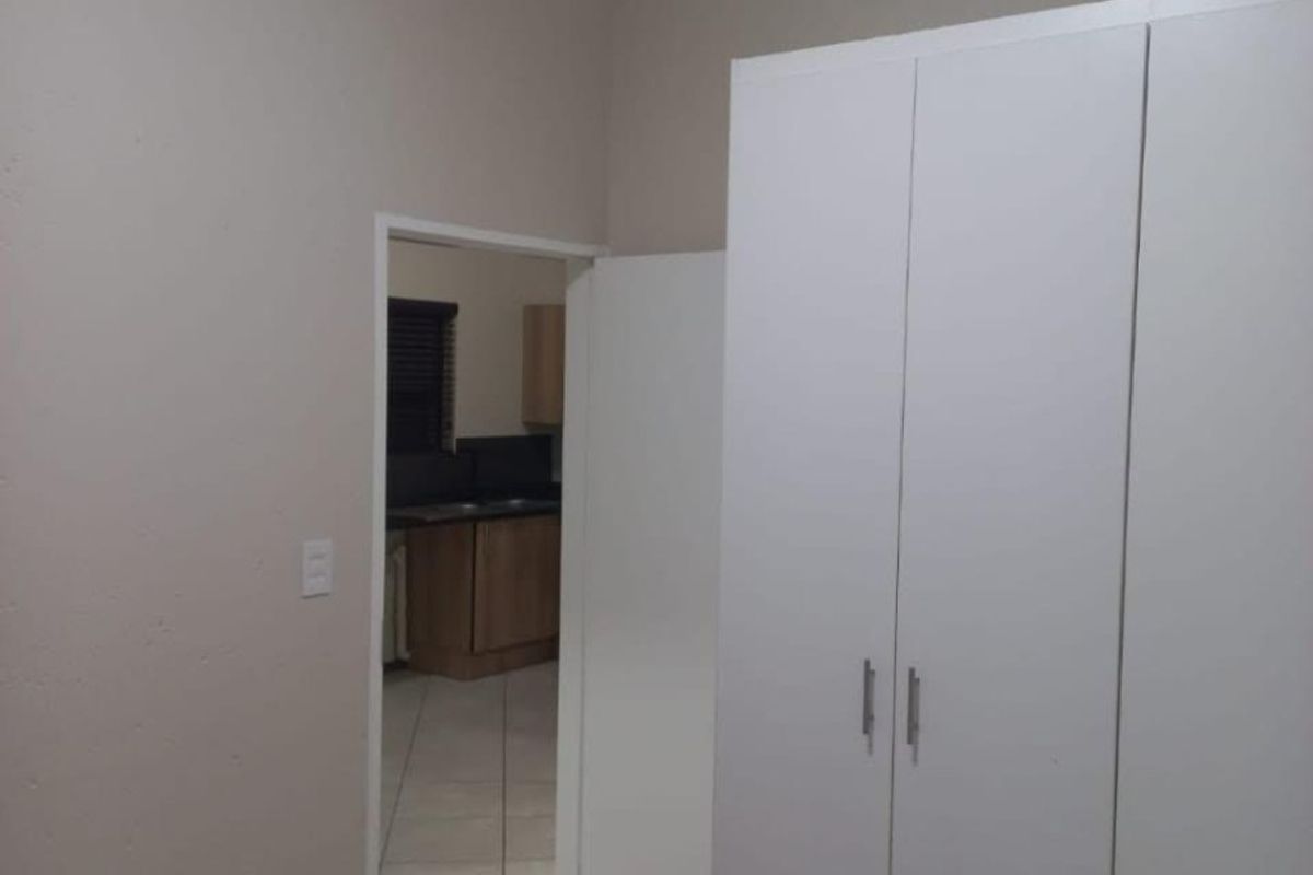 A clean, empty bedroom featuring neutral walls, tiled flooring, and built-in white cupboards, with a view through to the adjacent kitchen area.