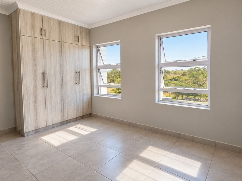 A bright, empty bedroom featuring neutral tiled flooring, built-in wooden cupboards, and large windows with views of the surrounding greenery.