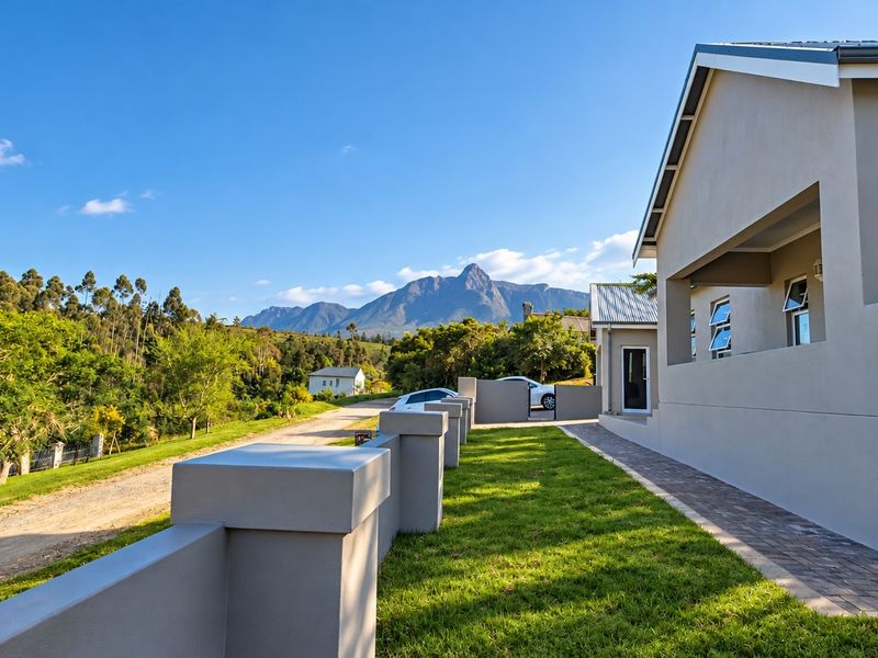 A well-maintained rear exterior view of the property featuring a paved walkway, manicured lawn, boundary wall, and scenic mountain views.