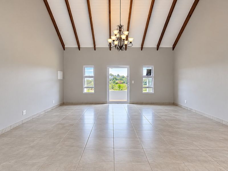 Spacious living area featuring a high vaulted ceiling with exposed timber beams, neutral tiled flooring, and a central chandelier, leading to an outdoor balcony.