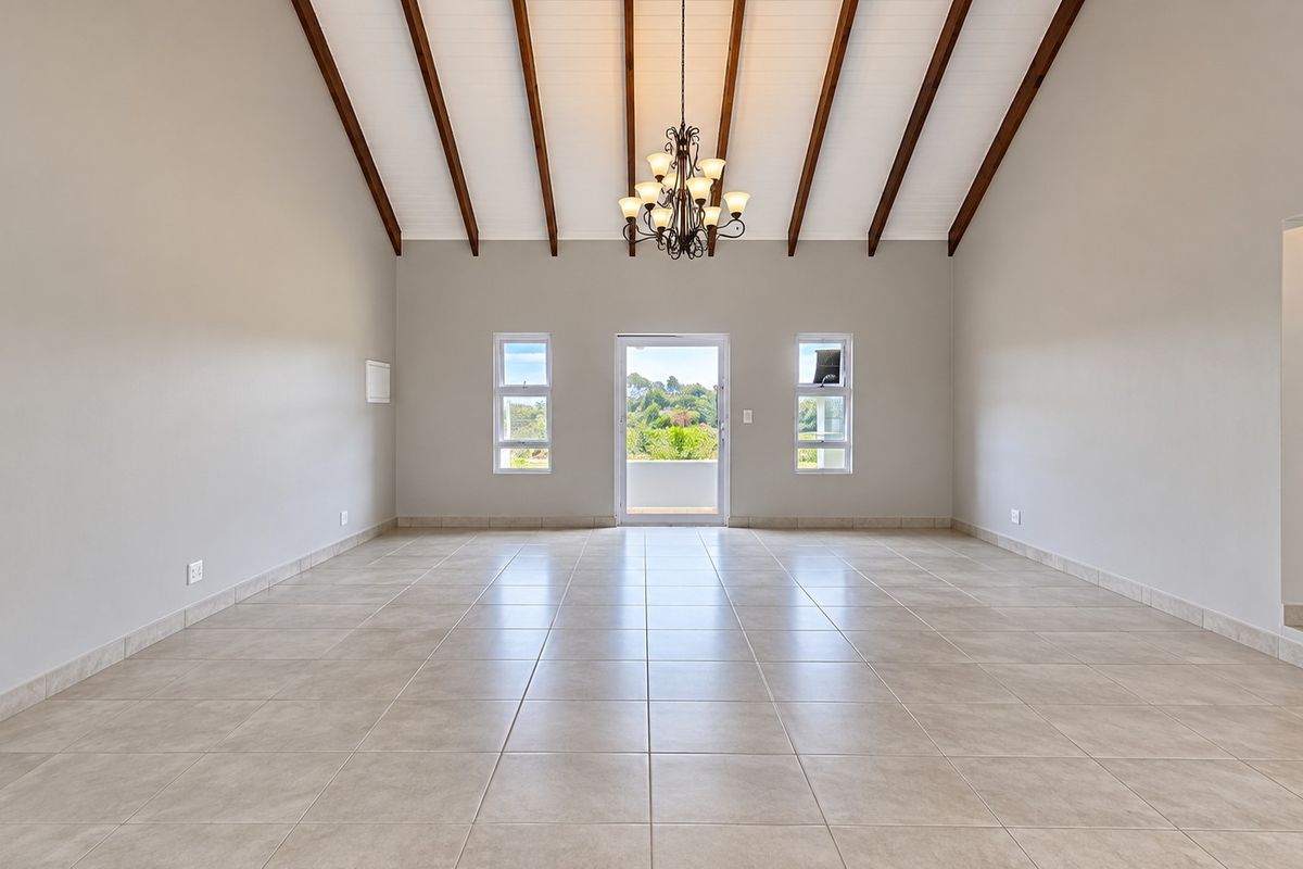 Spacious living area featuring a high vaulted ceiling with exposed timber beams, neutral tiled flooring, and a central chandelier, leading to an outdoor balcony.