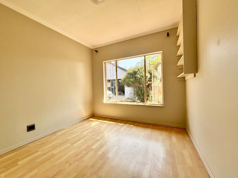 A bright, empty bedroom featuring light-toned laminate flooring, neutral walls, and built-in shelving, with a large window providing natural light and garden views.