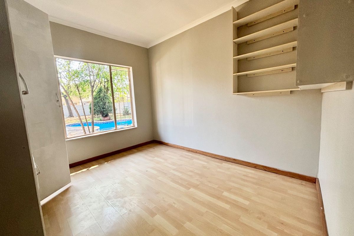 A bright, empty bedroom featuring light laminate flooring, built-in wall shelving, and a window view overlooking the garden and pool area.