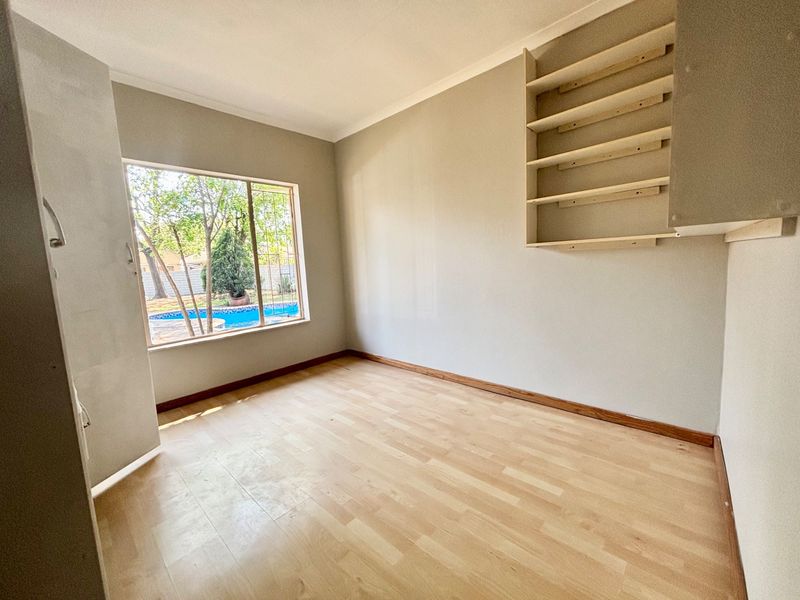 A bright, empty bedroom featuring light laminate flooring, built-in wall shelving, and a window view overlooking the garden and pool area.