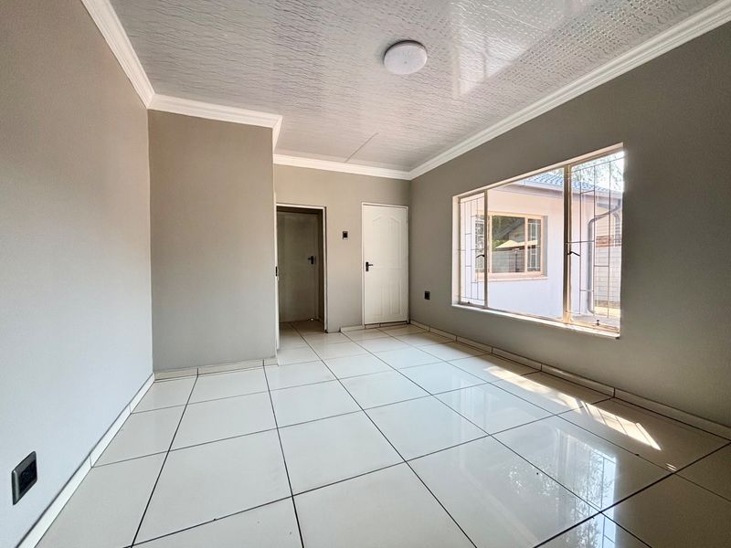 A bright, empty living room featuring neutral grey walls, large windows with security bars, and clean white tiled flooring throughout.