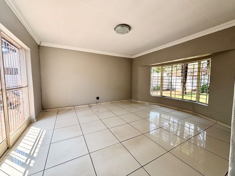 A spacious, empty living room featuring large tiled floors, a bay window with security bars, and a sliding door leading to the exterior.