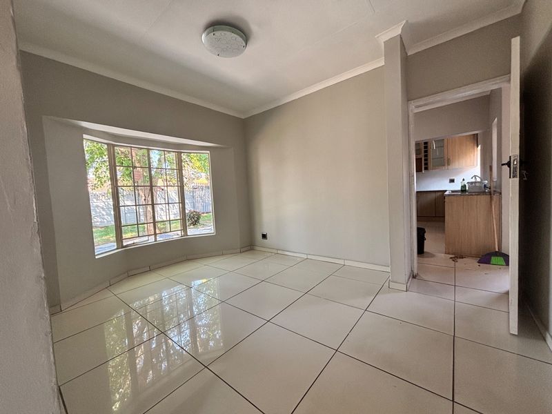 A bright, empty living room featuring large bay windows, neutral tiled flooring, and coved ceilings, with a view into the adjacent kitchen area.