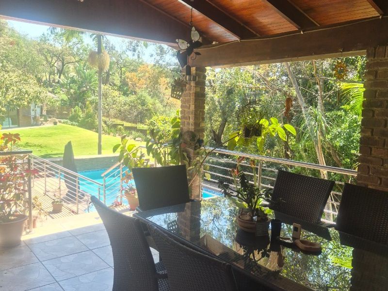A spacious covered patio featuring a timber-clad vaulted ceiling, exposed brick pillars, and tiled flooring, overlooking the garden and swimming pool area.