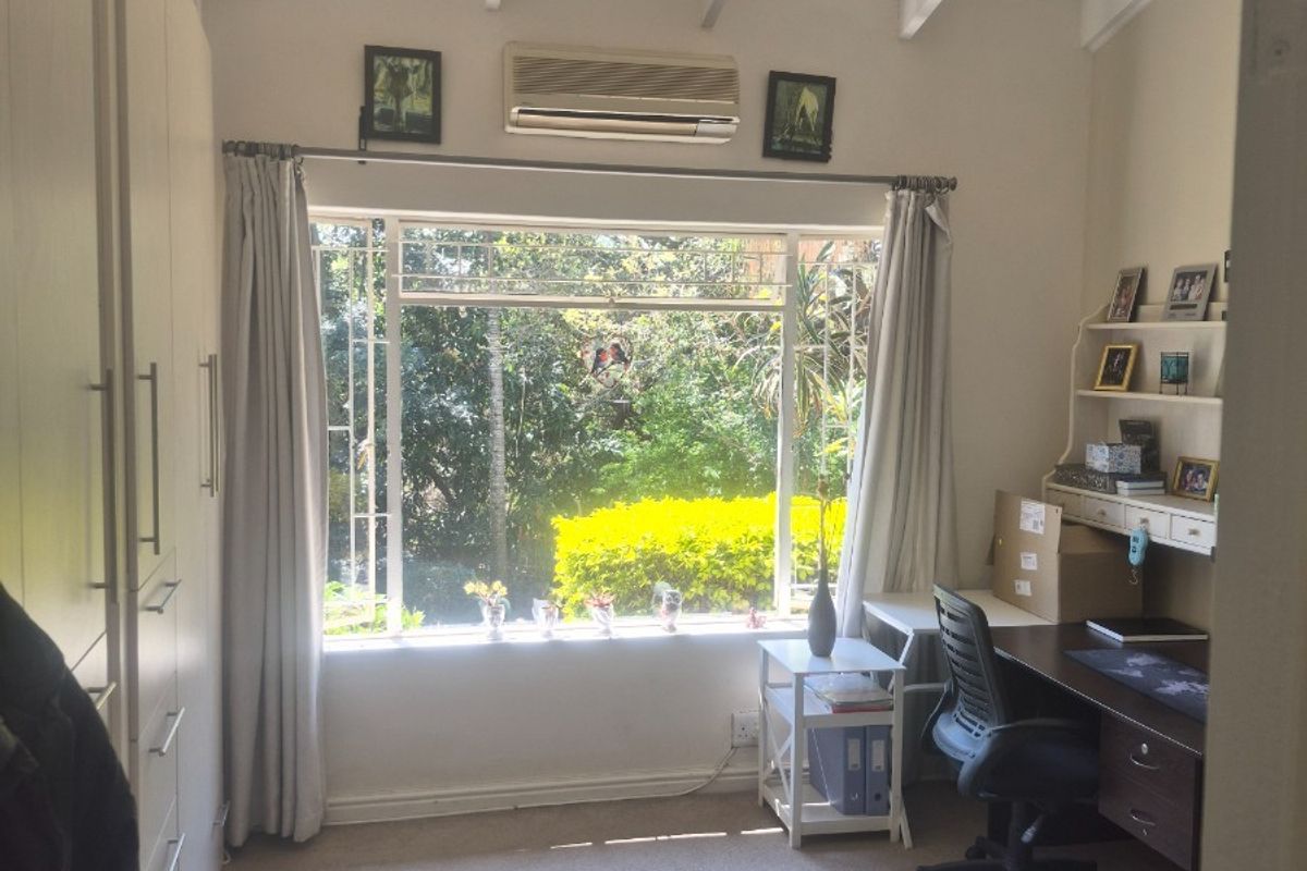 A bedroom featuring built-in cupboards, exposed ceiling beams, a split-system air conditioner, and a large window overlooking the garden.