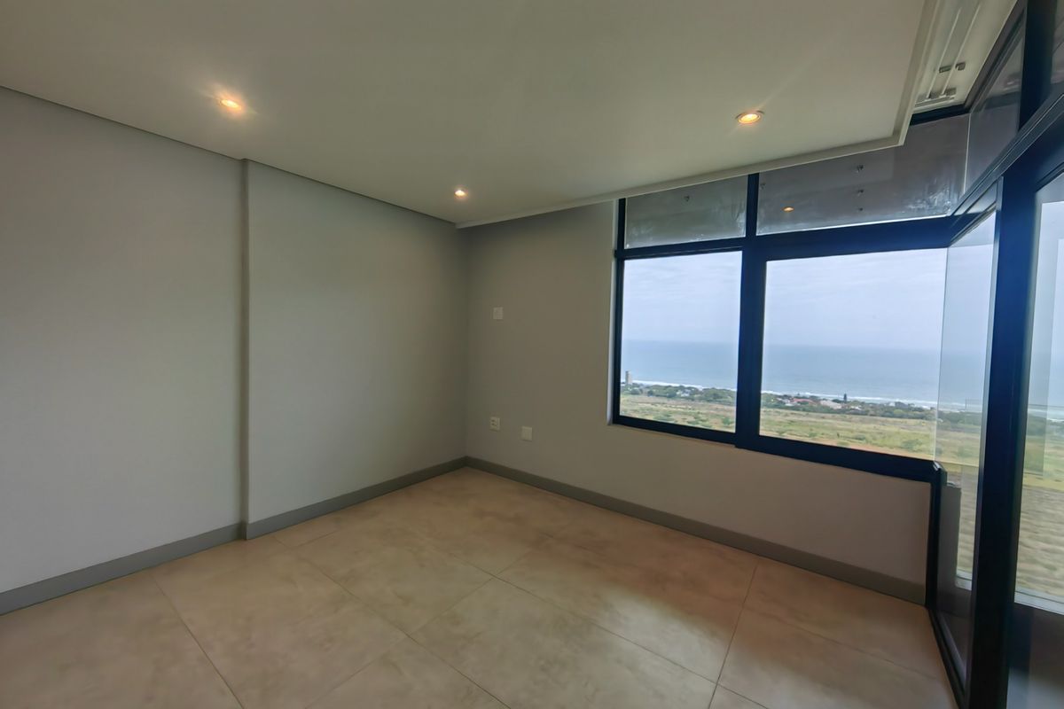 A bright, empty bedroom featuring large format floor tiles, recessed lighting, and expansive windows offering a scenic coastal view.