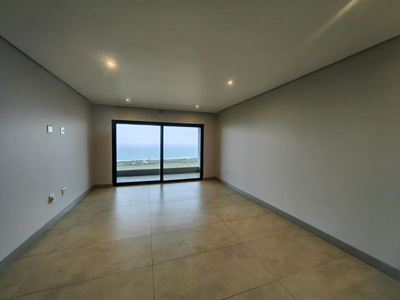 A bright, empty living area featuring large sliding glass doors that open to a scenic coastal view, complemented by modern tiled flooring and recessed lighting.