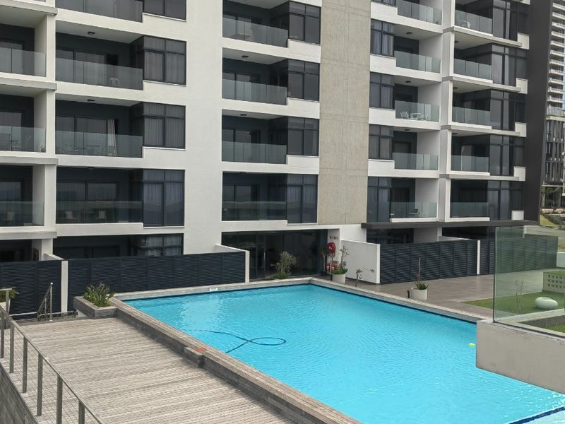 A well-maintained outdoor swimming pool area featuring timber decking and glass balustrades, set against a modern multi-story apartment building with private balconies.