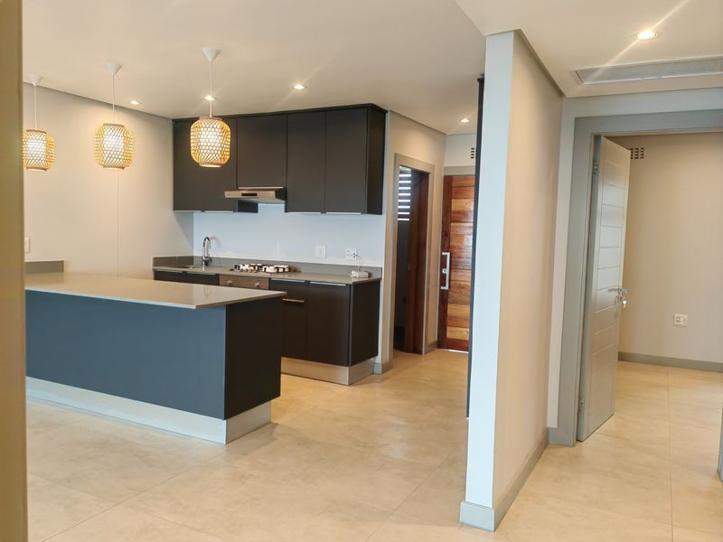 Modern open-plan kitchen featuring dark cabinetry, stone countertops, gas hob, and integrated extractor, flowing into a bright, tiled living area with recessed lighting.
