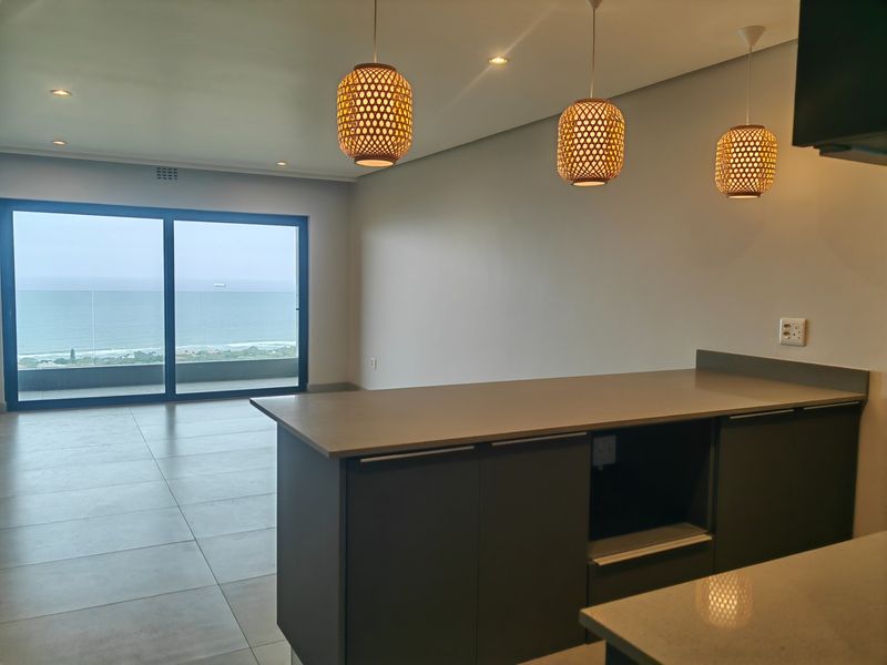 Modern open-plan living area featuring a sleek kitchen island, tiled floors, and large sliding doors opening to a balcony with expansive ocean views.