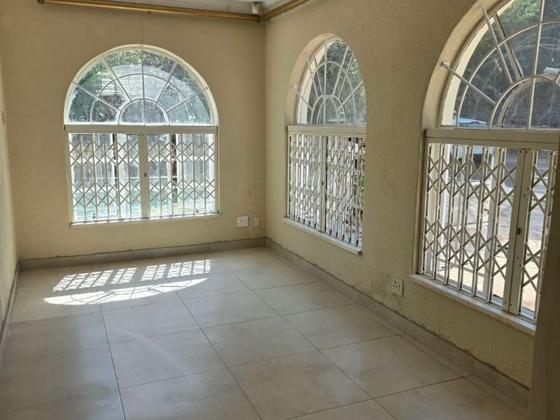 An empty living area featuring arched windows with security gates, tiled flooring, and neutral wall finishes, offering a bright and versatile space.