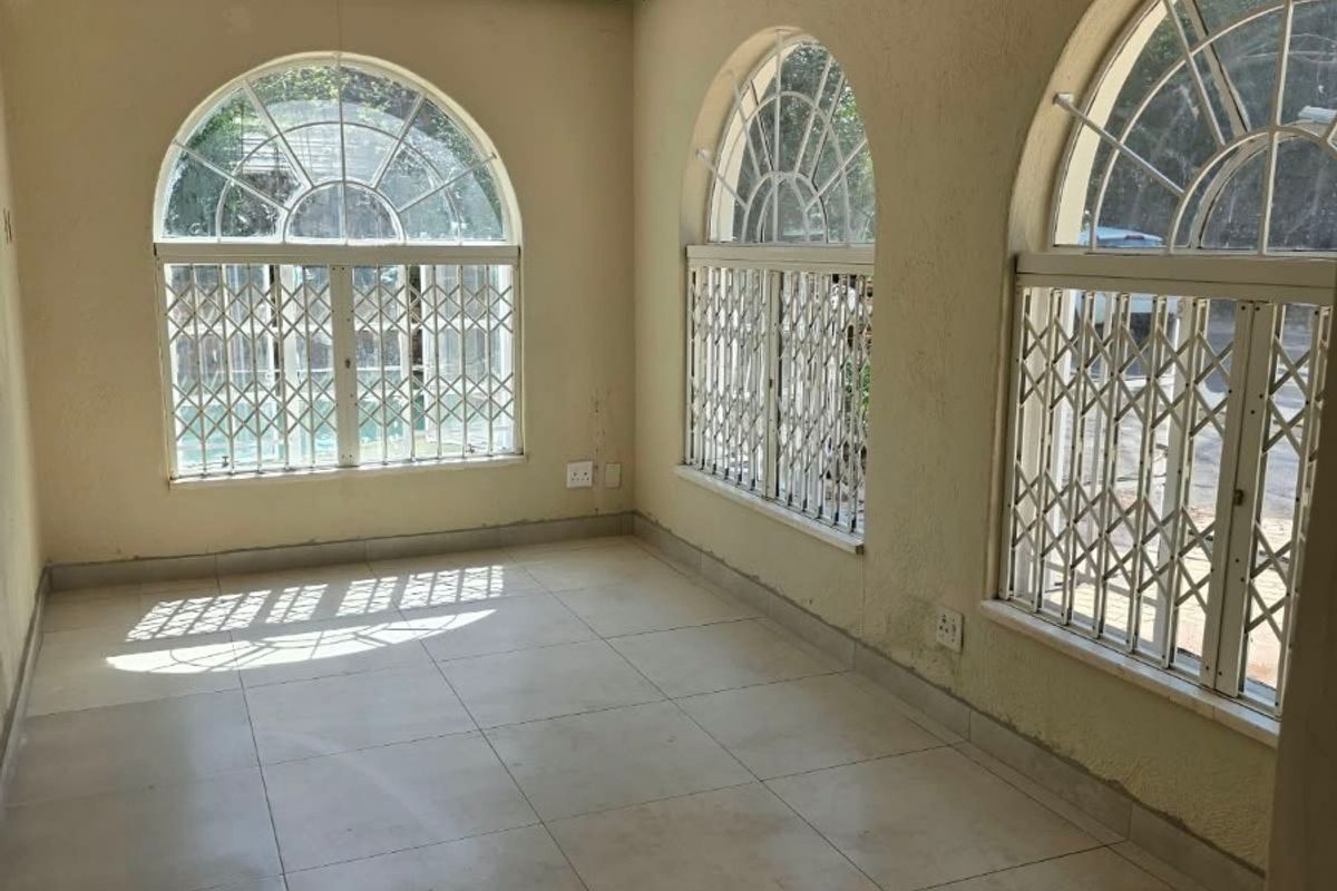 An empty living area featuring arched windows with security gates, tiled flooring, and neutral wall finishes, offering a bright and versatile space.