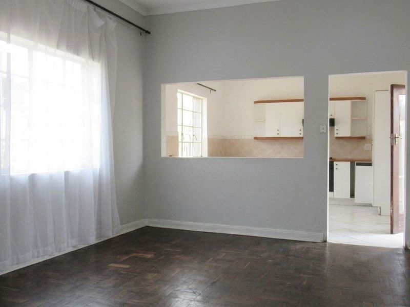 A bright, empty living room featuring classic parquet flooring, a convenient serving hatch to the kitchen, and neutral wall finishes.
