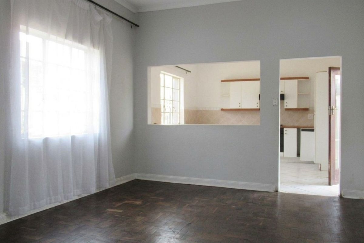 A bright, empty living room featuring classic parquet flooring, a convenient serving hatch to the kitchen, and neutral wall finishes.