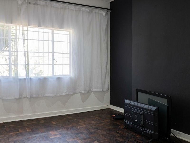 A bright living room featuring dark parquet flooring and a large window with security bars, finished with a bold black accent wall.