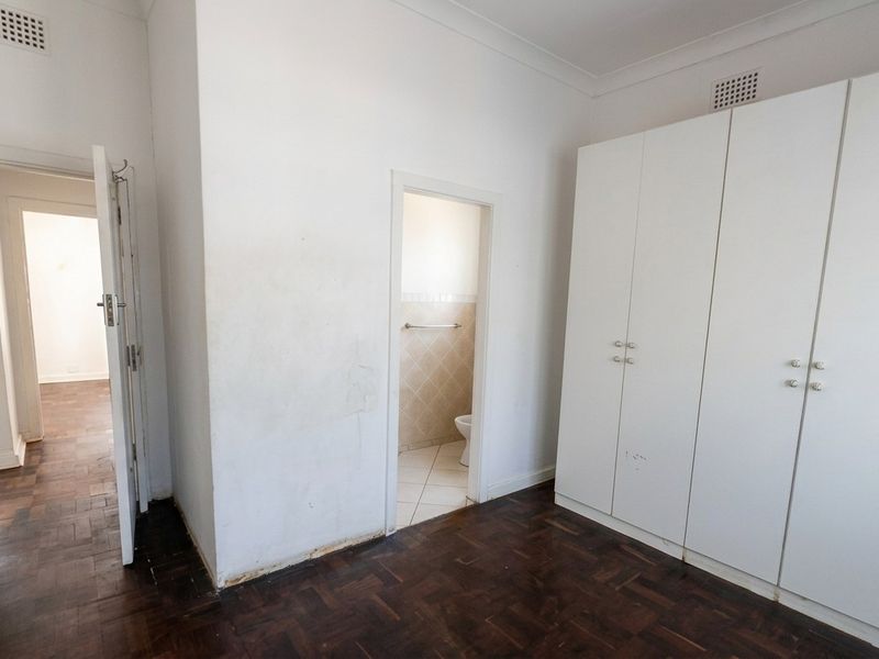 An empty bedroom featuring dark parquet flooring, built-in white cupboards, and an en-suite bathroom access point.