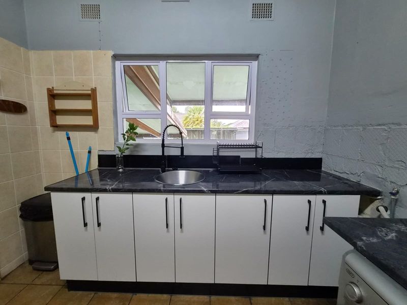 A functional scullery area featuring modern white cabinetry, a dark marble-effect countertop, stainless steel sink, and a window providing natural light.
