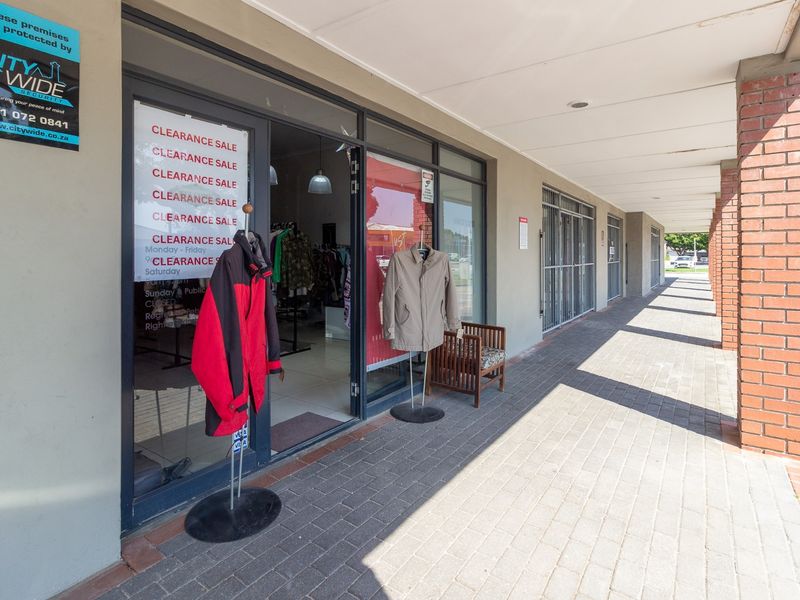 Front exterior view of a retail storefront featuring a paved walkway, brick columns, and glass entrance doors with security gates.