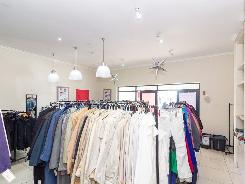 A retail shop interior featuring tiled flooring, pendant lighting, and a glass entrance door, currently utilized for clothing display.