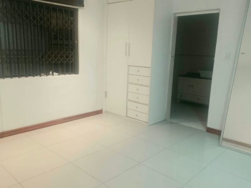 A clean, empty bedroom featuring white tiled flooring, built-in cupboards, and a window fitted with a security gate.