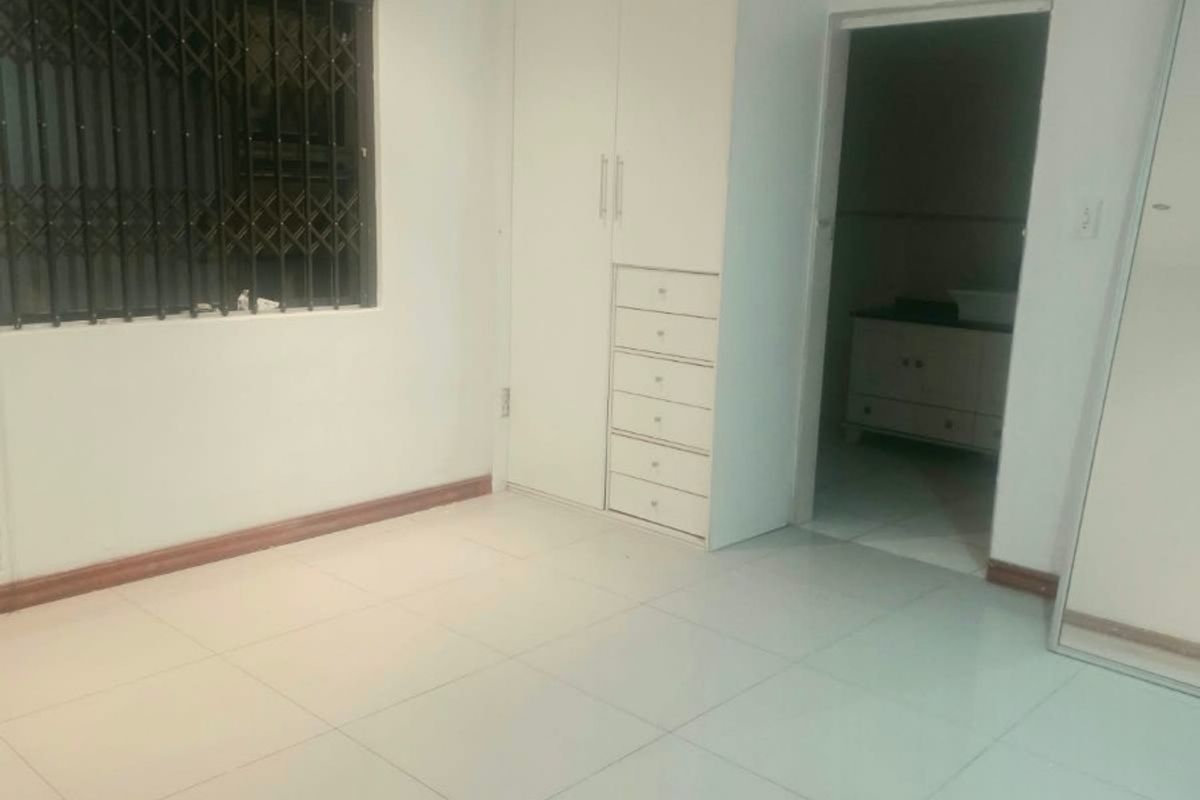 A clean, empty bedroom featuring white tiled flooring, built-in cupboards, and a window fitted with a security gate.