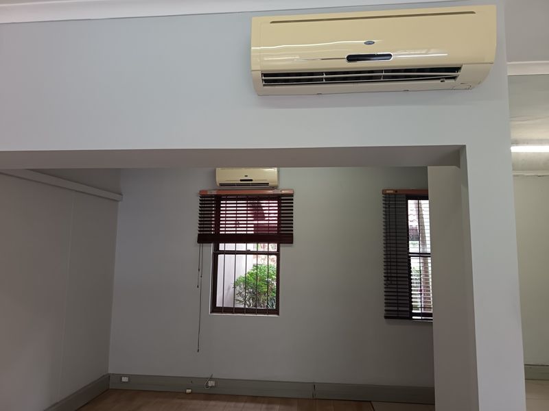 A bright, empty office space featuring dual split-system air conditioning units, timber venetian blinds, and security bars on the windows.