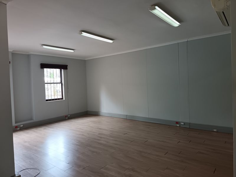 A spacious, empty office area featuring laminate flooring, neutral walls, fluorescent lighting, and an integrated air conditioning unit.
