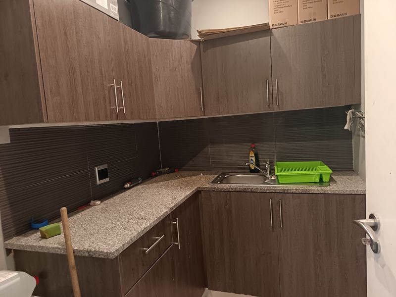 A compact kitchenette featuring dark wood-grain cabinetry, granite countertops, a stainless steel sink, and a modern textured tiled backsplash.