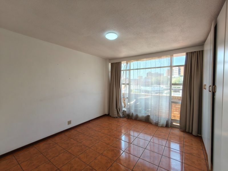 A bright, empty bedroom featuring tiled flooring, built-in cupboards, and a large window providing natural light and an urban view.