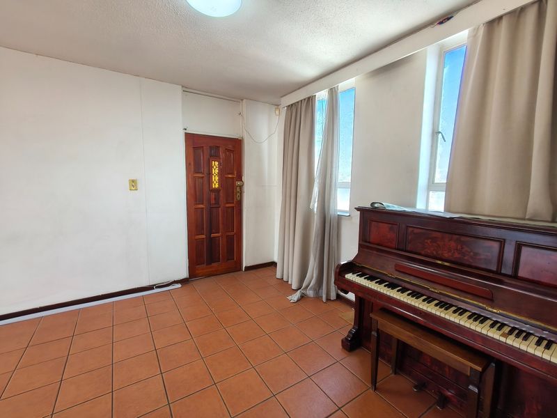 A living area featuring terracotta-style tiled flooring, a solid timber entrance door, and large windows allowing natural light into the space.