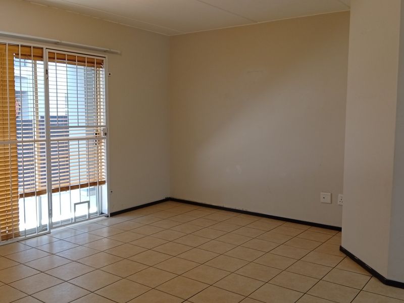 An empty living area featuring neutral tiled flooring, plain walls, and a sliding glass door with integrated security gate and blinds.