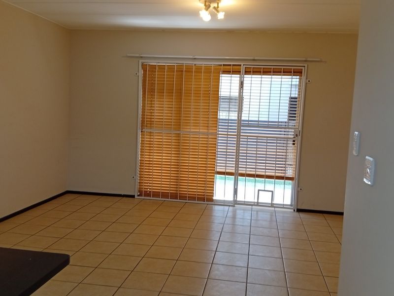 A spacious, empty living area featuring neutral tiled flooring and a sliding glass door with security gate and blinds leading to the exterior.