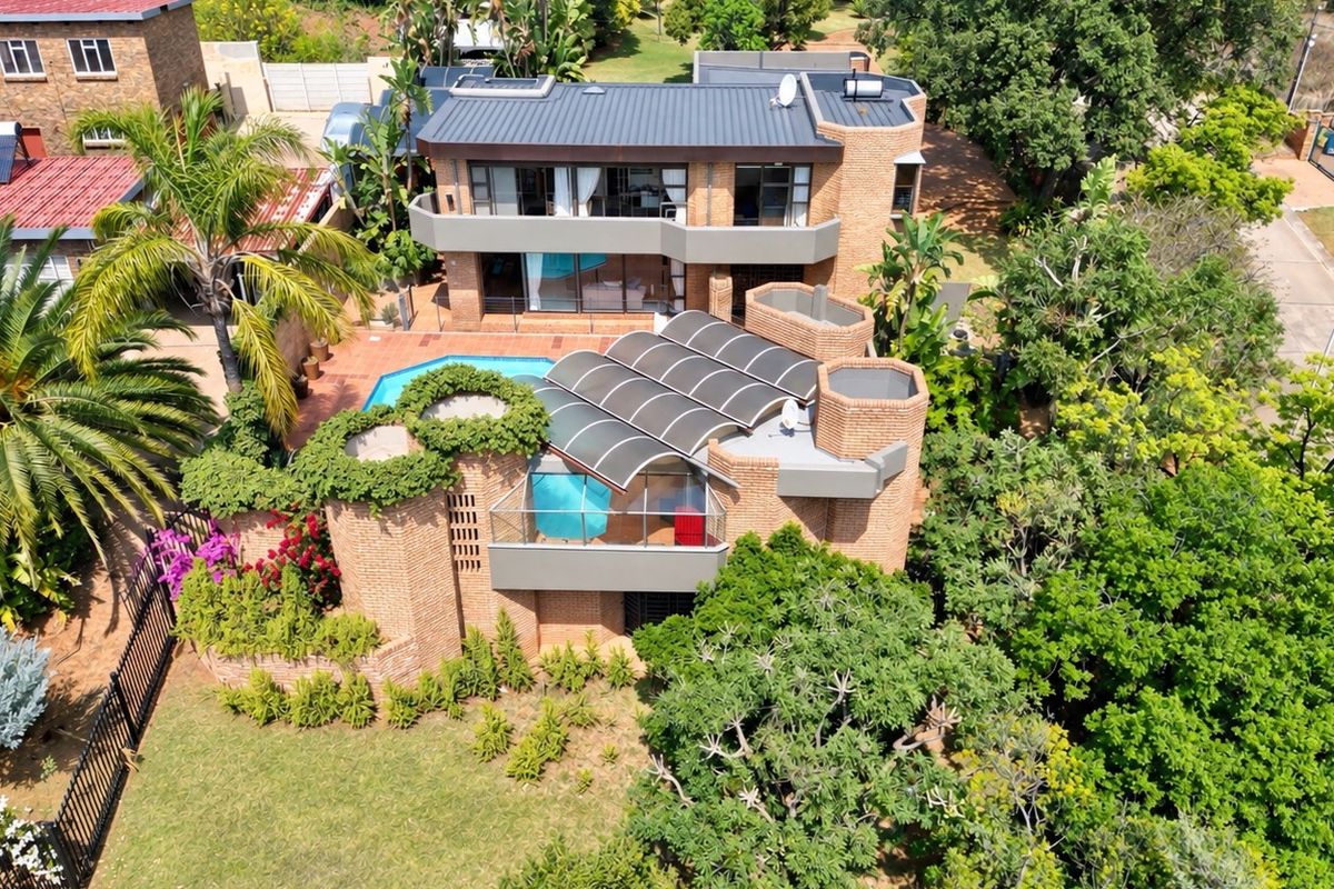 Aerial view of a multi-level brick residence featuring a swimming pool, expansive balconies, and a unique curved patio roof structure set in a lush garden.