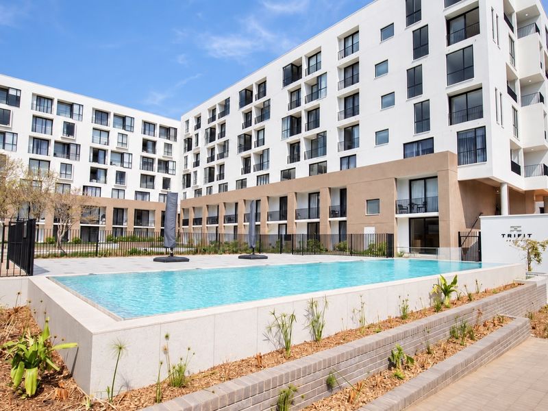 A modern apartment complex featuring a clean, well-maintained swimming pool area with paved decking, stone coping, and secure perimeter fencing.