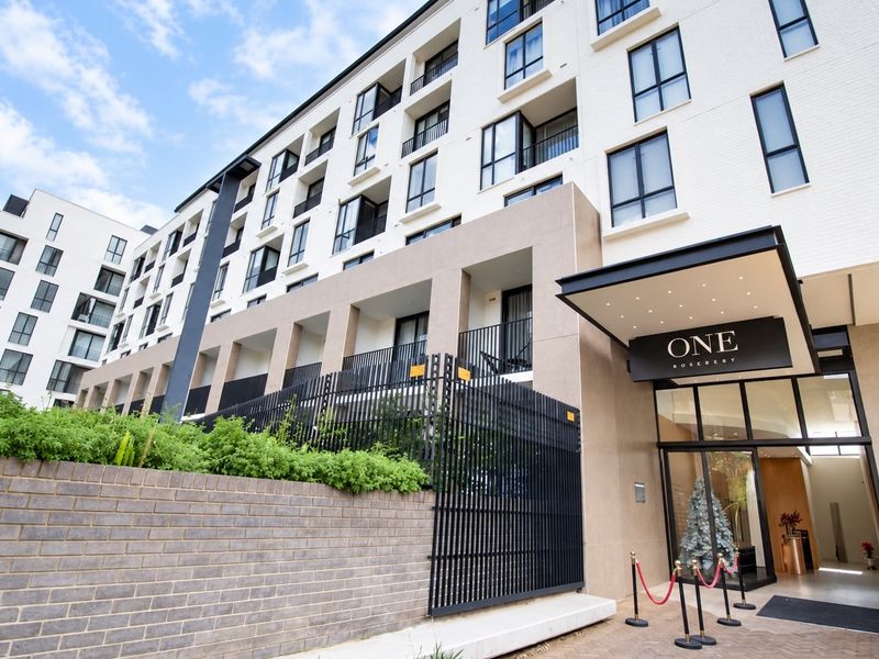 Modern apartment building exterior featuring a brick facade, secure metal fencing, and a prominent entrance canopy with recessed lighting.