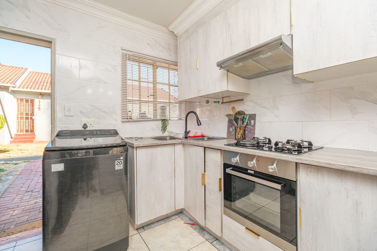 Compact kitchen featuring light wood-look cabinetry, gas hob, built-in oven, extractor fan, and marble-effect wall tiling with direct access to the exterior.